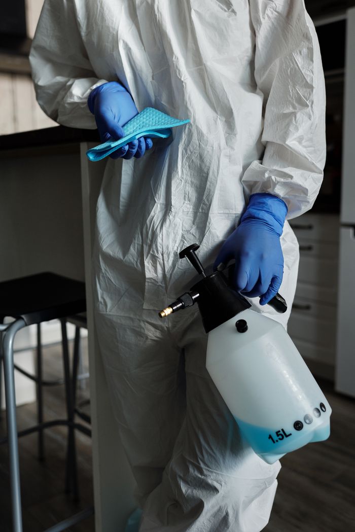 Individual in full protective gear holding disinfectant spray and gloves indoors for safety.