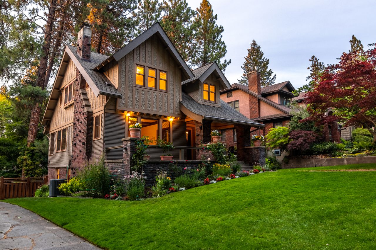 A beautifully lit Victorian-style house surrounded by a lush garden at twilight, showcasing classic architecture.