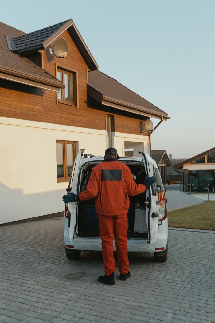 A delivery man in red uniform unloading a van in a residential driveway during the day.